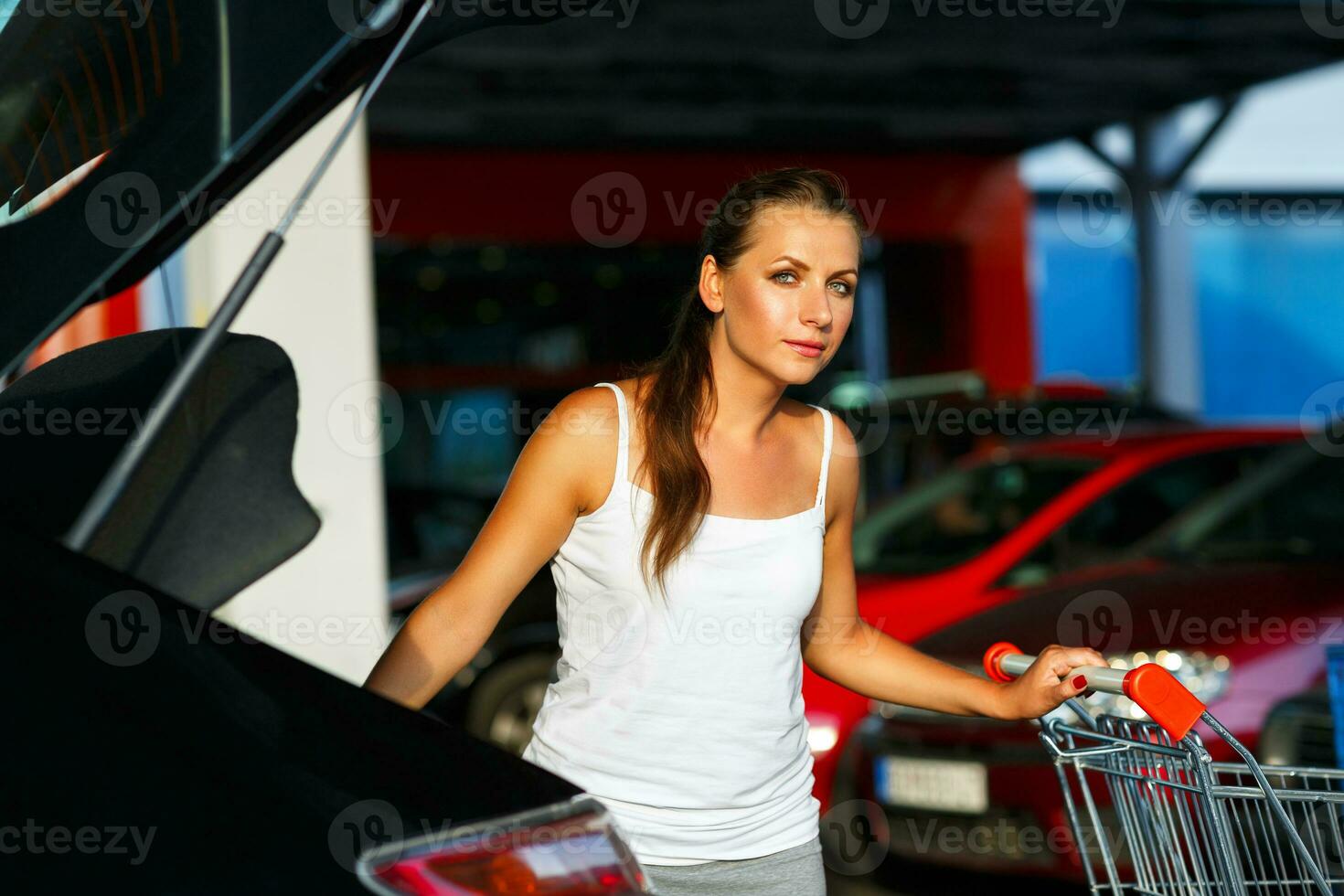 ung kvinna skift de inköp från handla vagn i de trunk av en bil på de parkering foto