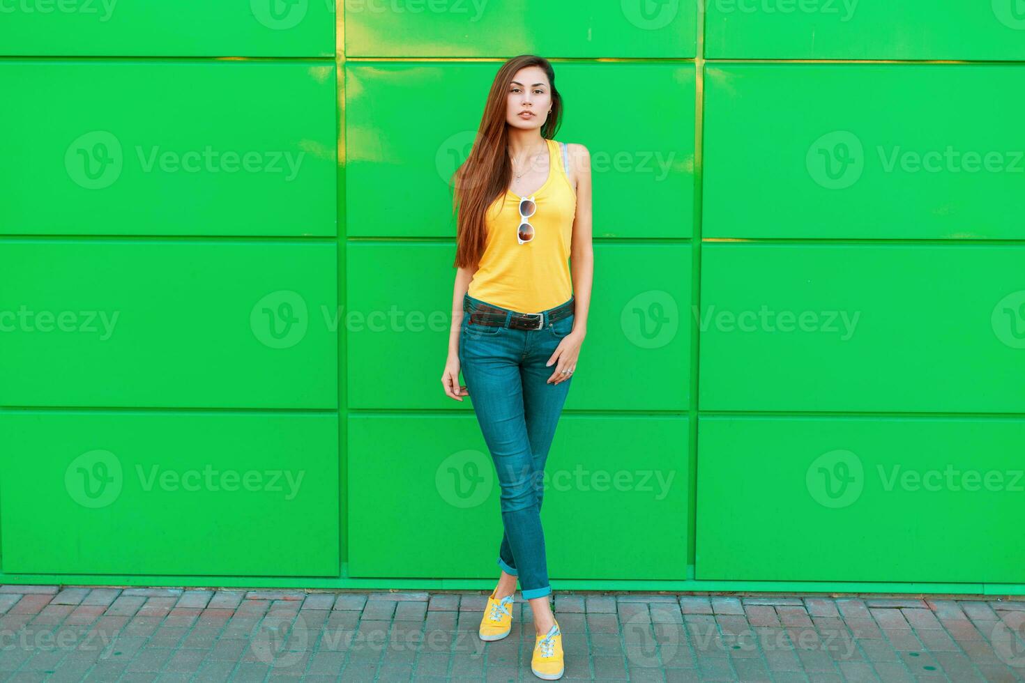 modern skön kvinna i eleganta gul kläder och jeans stående nära en grön metall vägg. foto
