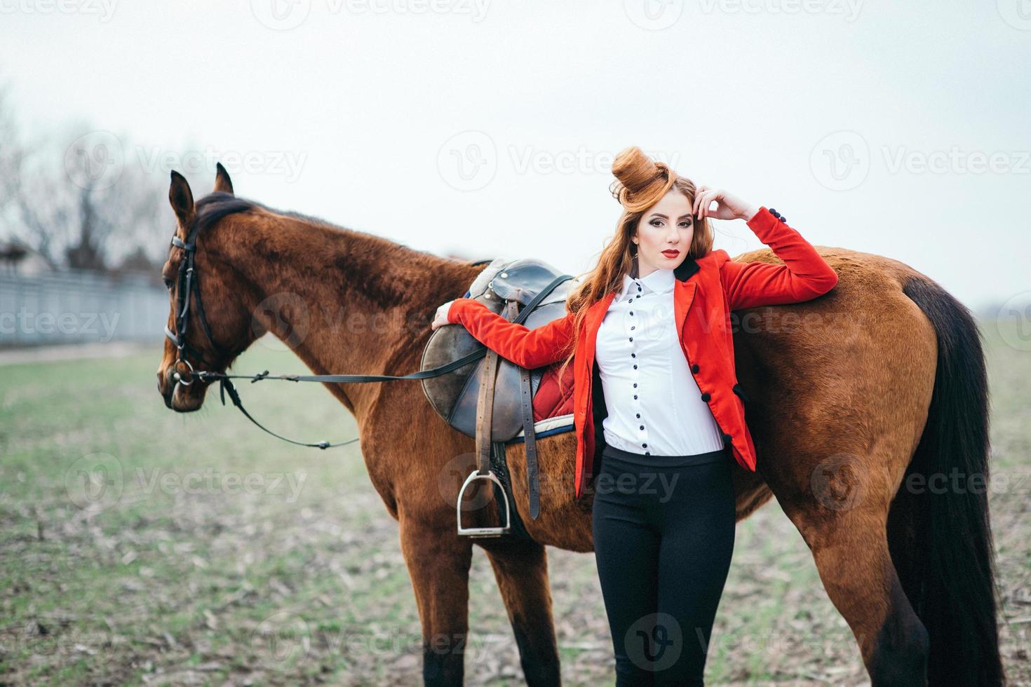 rödhårig jockeytjej i en röd kofta och svarta höga stövlar med en häst foto