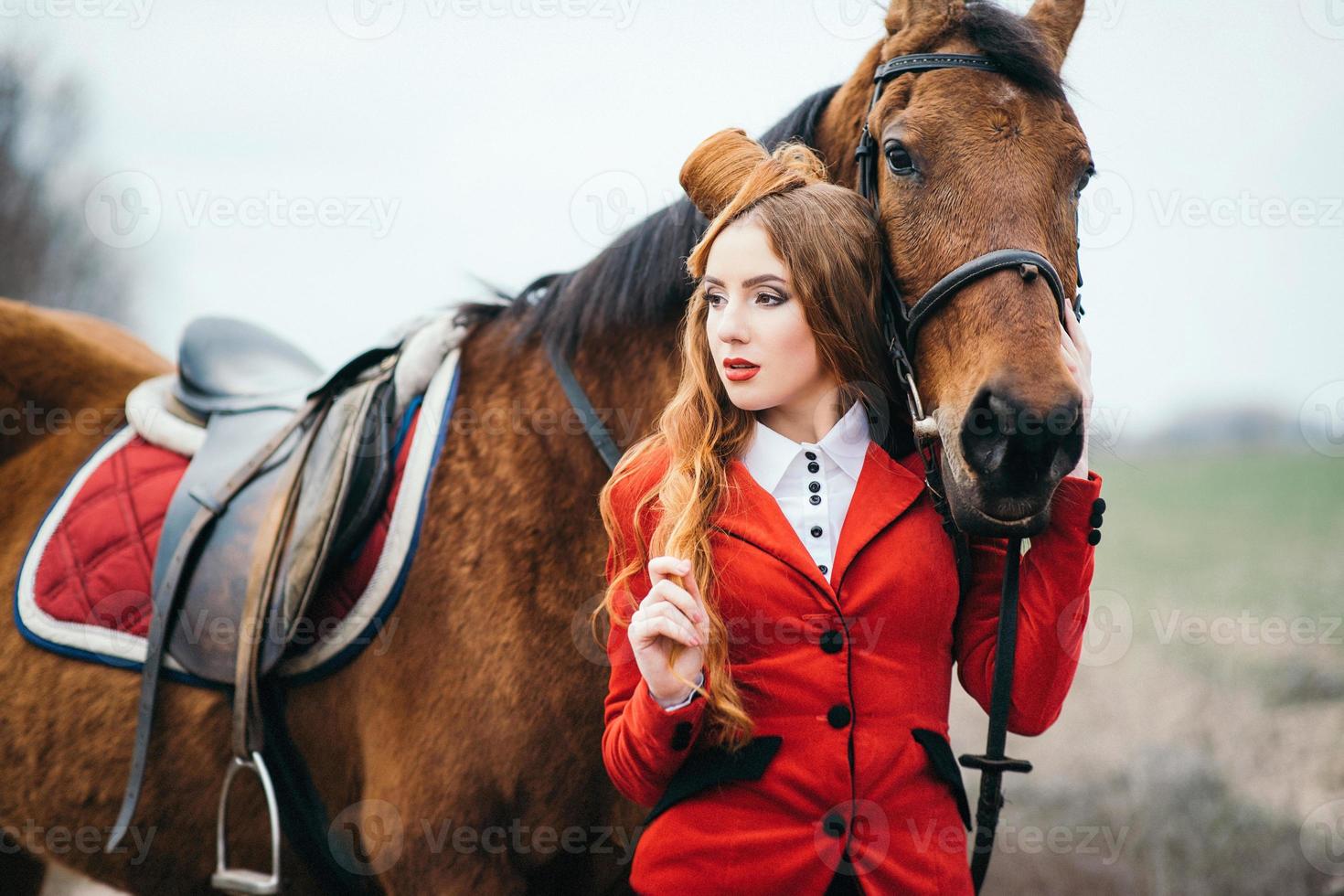 rödhårig jockeytjej i en röd kofta och svarta höga stövlar med en häst foto