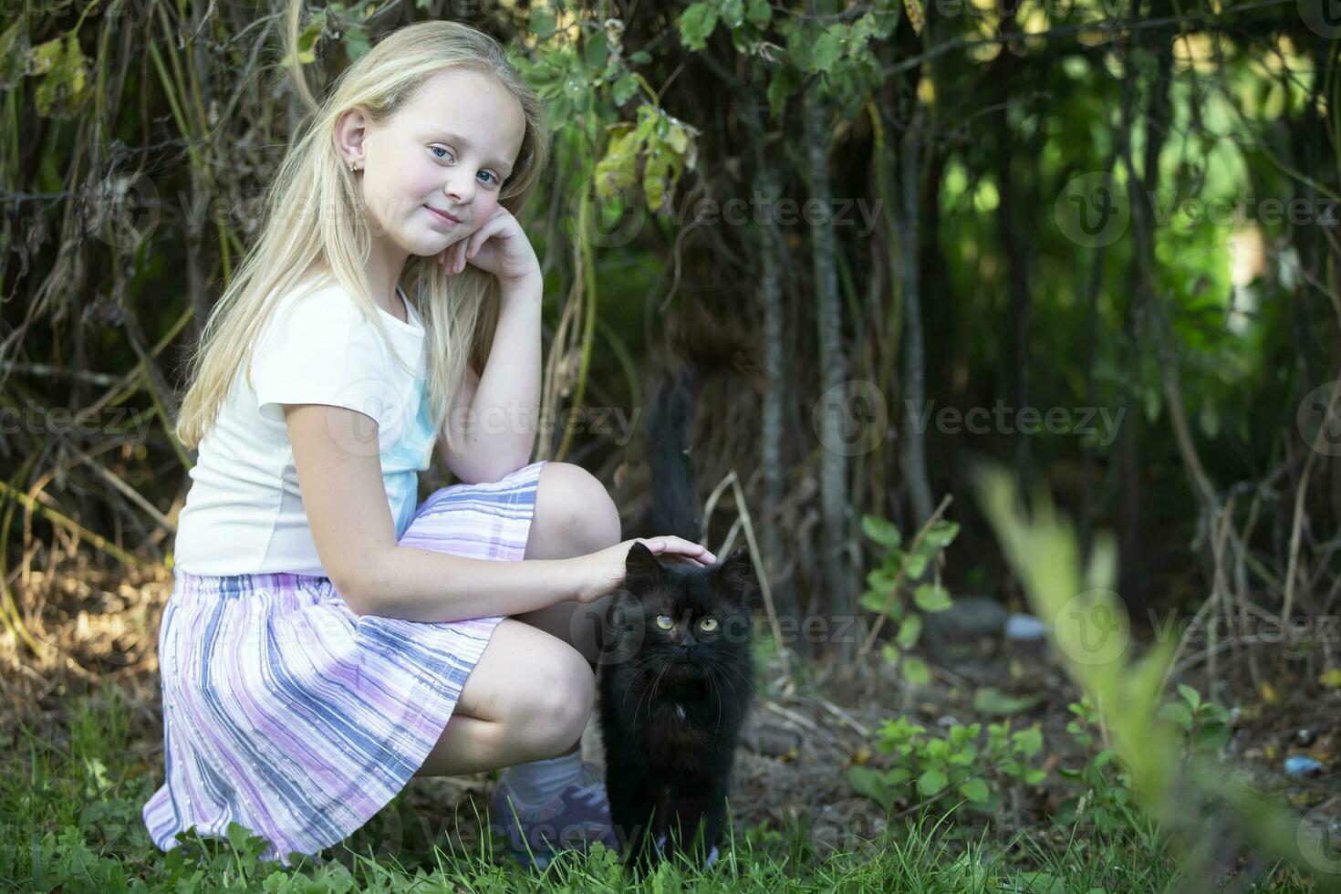 liten blond flicka spelar med en svart katt på en bakgrund av grön buskar. foto