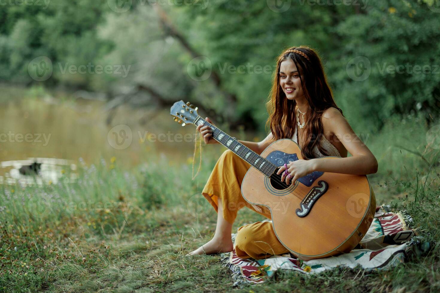 ung hippie kvinna Sammanträde utomhus på en pläd och spelar henne gitarr på de flodbank in i de solnedgång foto