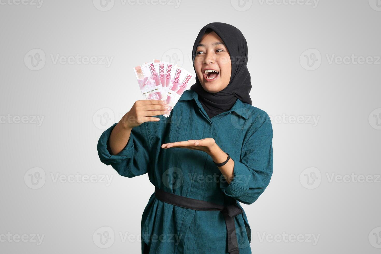 porträtt av upphetsad asiatisk hijab kvinna i tillfällig utrusta som visar och pekande ett hundra tusen rupiah. finansiell och besparingar begrepp. isolerat bild på vit bakgrund foto