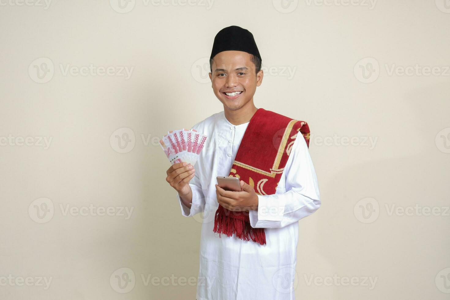 porträtt av attraktiv asiatisk muslim man i vit skjorta som visar ett hundra tusen rupiah medan använder sig av mobil telefon. finansiell och besparingar begrepp. isolerat bild på grå bakgrund foto