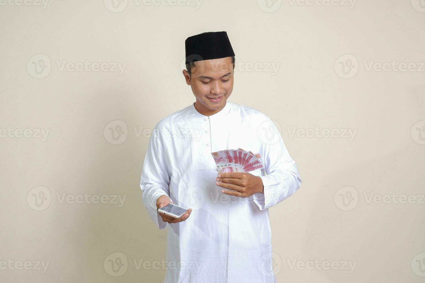 porträtt av attraktiv asiatisk muslim man i vit skjorta som visar ett hundra tusen rupiah medan använder sig av mobil telefon. finansiell och besparingar begrepp. isolerat bild på grå bakgrund foto