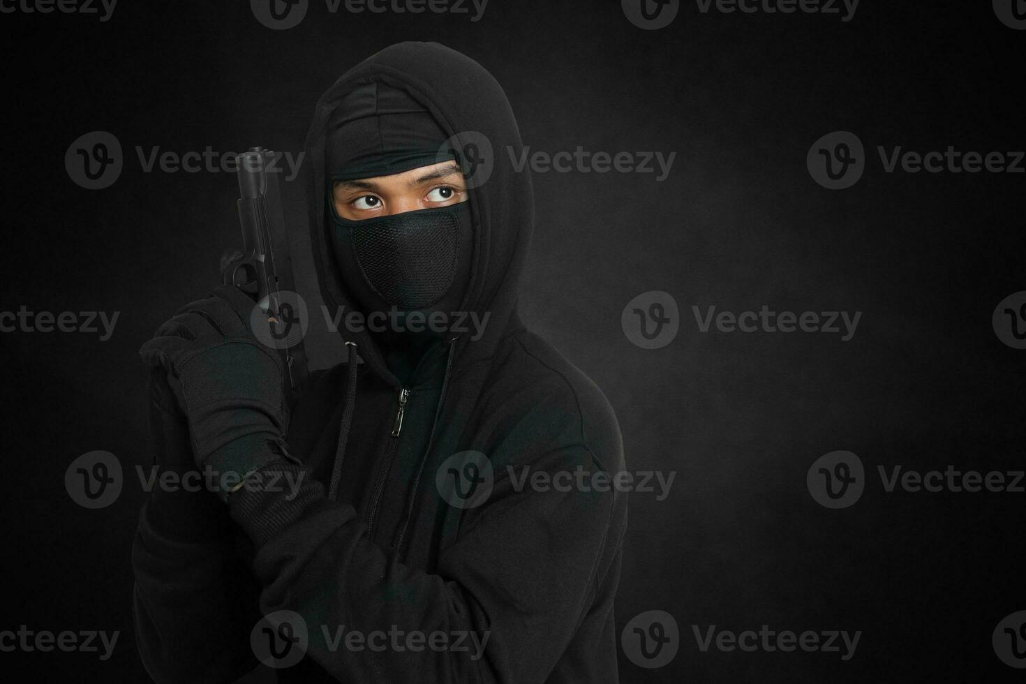 mystisk man bär svart luvtröja och mask innehav en pistol, skytte med en pistol. isolerat bild på mörk omgivande bakgrund foto