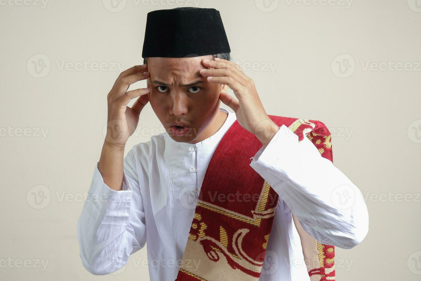 porträtt av attraktiv asiatisk muslim man i vit skjorta med kalott känsla stilig och självsäker. rörande hans tempel till göra en stil. isolerat bild på grå bakgrund foto