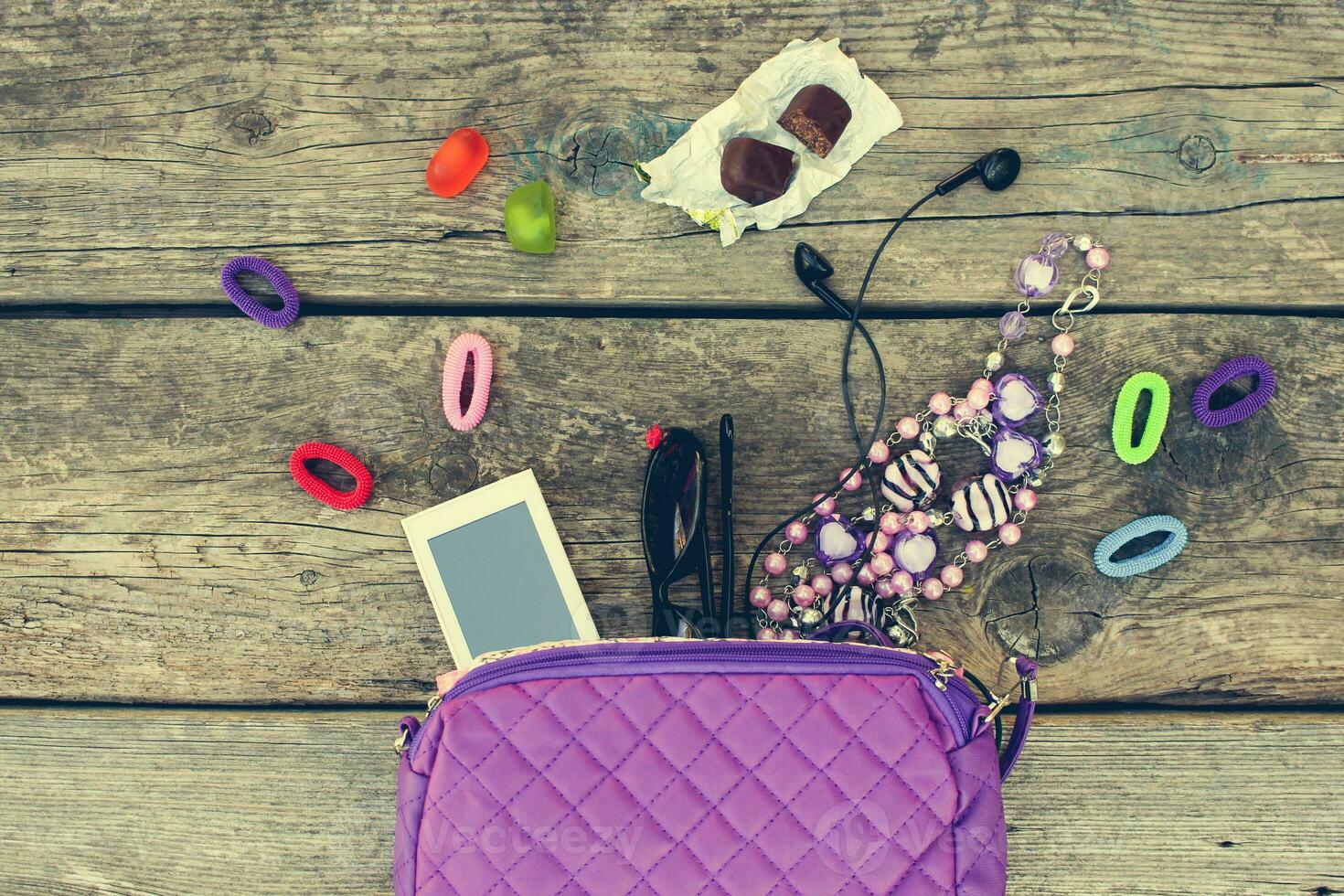 barns handväska och Tillbehör mobil telefon, hår band, godis, pärlor, hörlurar, solglasögon på gammal trä- bakgrund. topp se. tonad bild. foto