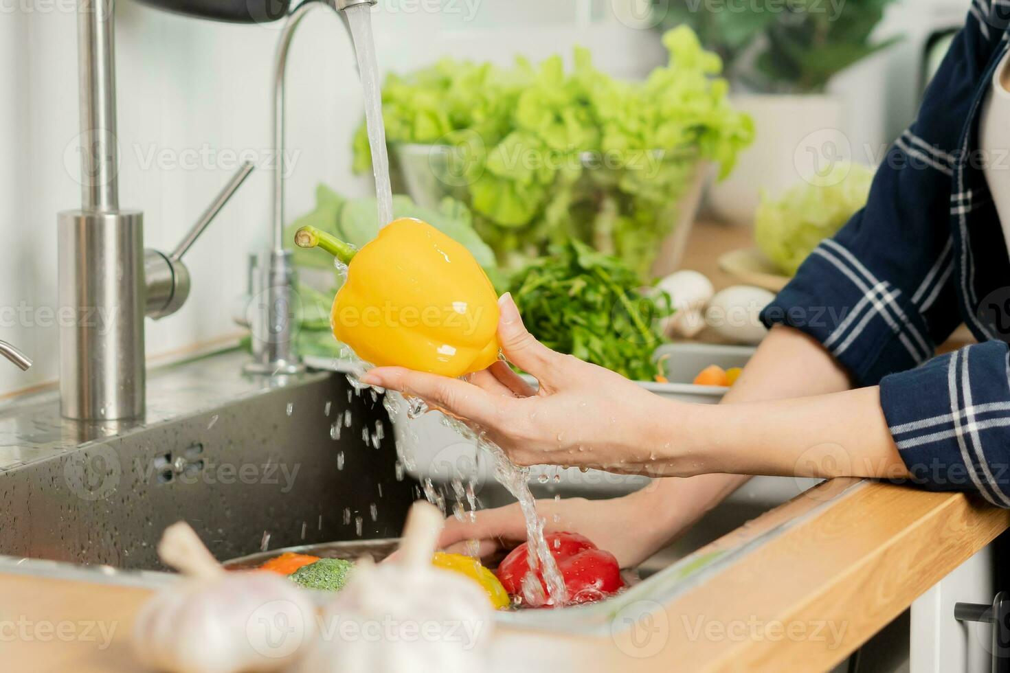 stänga upp hand av asiatisk ung hushållerska kvinna, tvättning ljuv peppar, gul paprika, grönsaker med stänk vatten i handfat av vatten på handfat i kök, framställning färsk sallad, matlagning måltid.hälsa mat foto