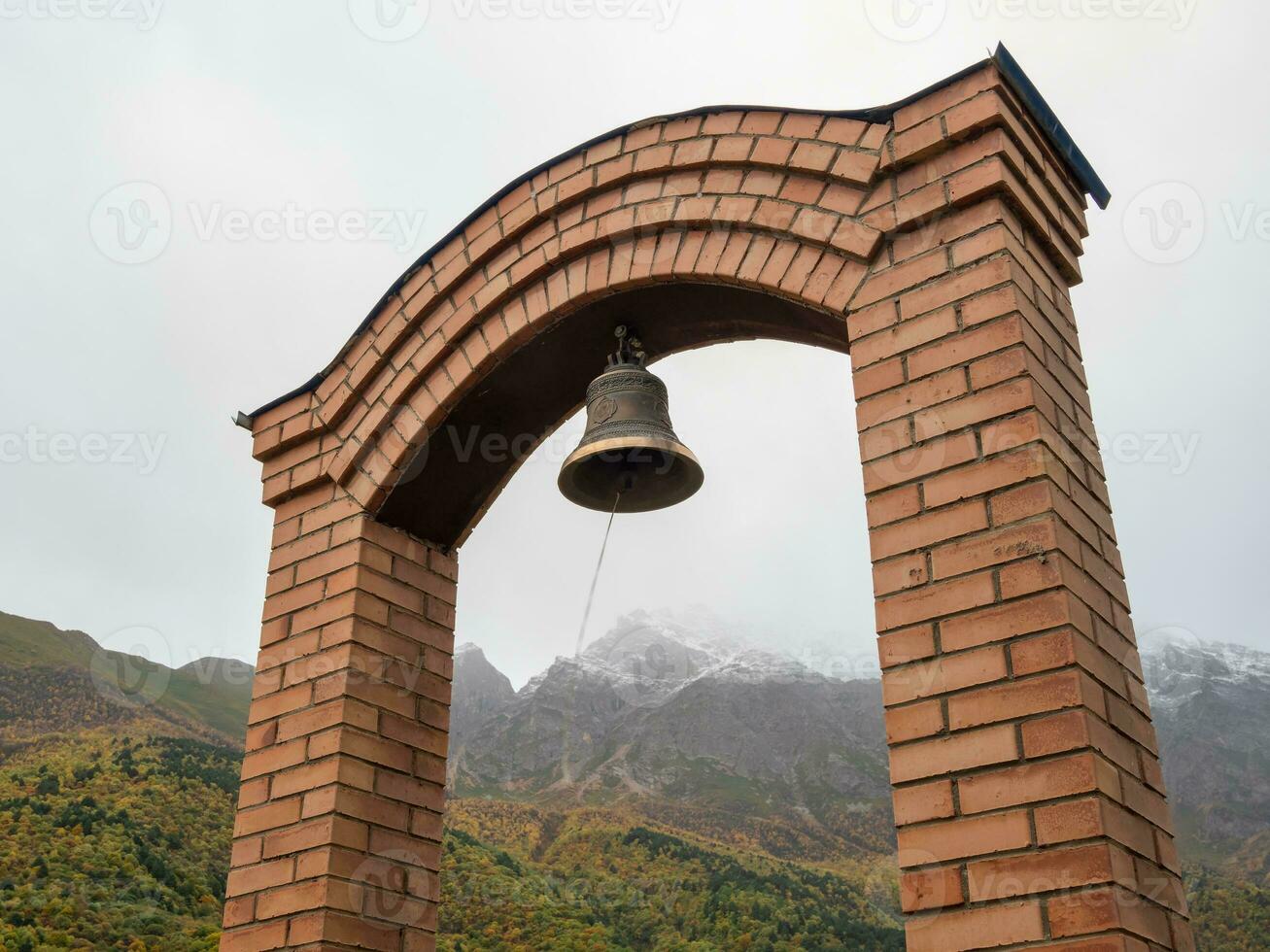 stock Foto av gammal koppar eller brons klocka hängande på en sten ram på kapell i hedra av de död- på dimmig berg bakgrund. bild fångad på irafsky distrikt, stur-digora by. Ryssland.