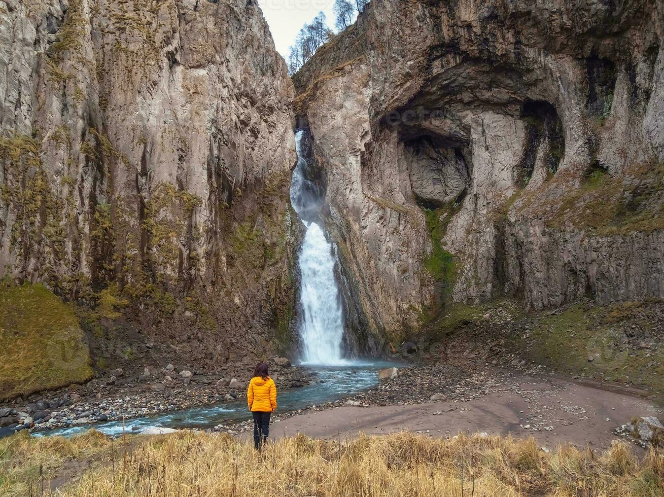 flicka i vattentät gul Kläder stanna kvar på de klippa på bakgrund av gil-su vattenfall i norr Kaukasus. se från tillbaka. foto