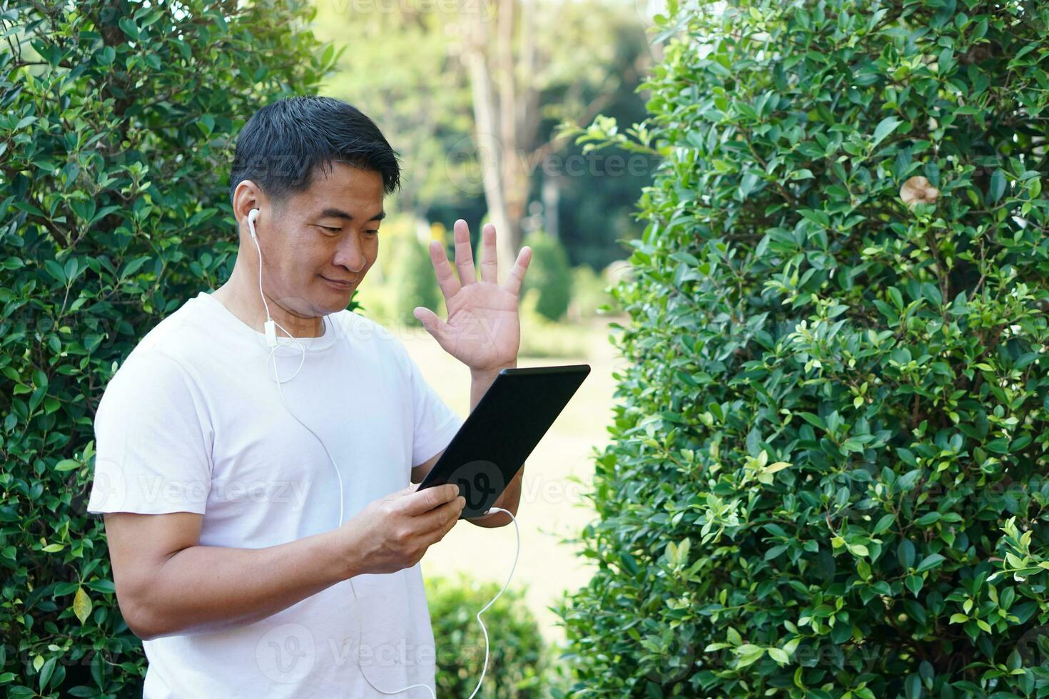 asiatisk man är framställning video ring upp och chattar genom smart läsplatta på de parkera. vinka hand. begrepp, koppla av tid. använder sig av trådlös teknologi internet till conect människor tillsammans.använda konferens möte app. foto