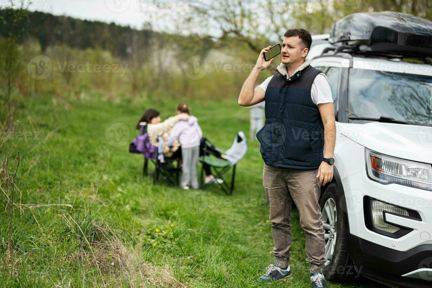 man i ärmlös väst tala på telefon mot sUV bil med tak kuggstång låda, familj på picknick. foto