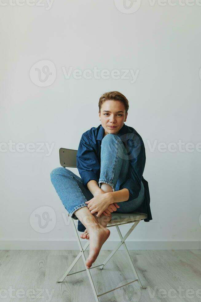 en ung kvinna Sammanträde i en stol på Hem leende med tänder med en kort frisyr i jeans och en denim skjorta på en vit bakgrund. flicka naturlig poser med Nej filter foto