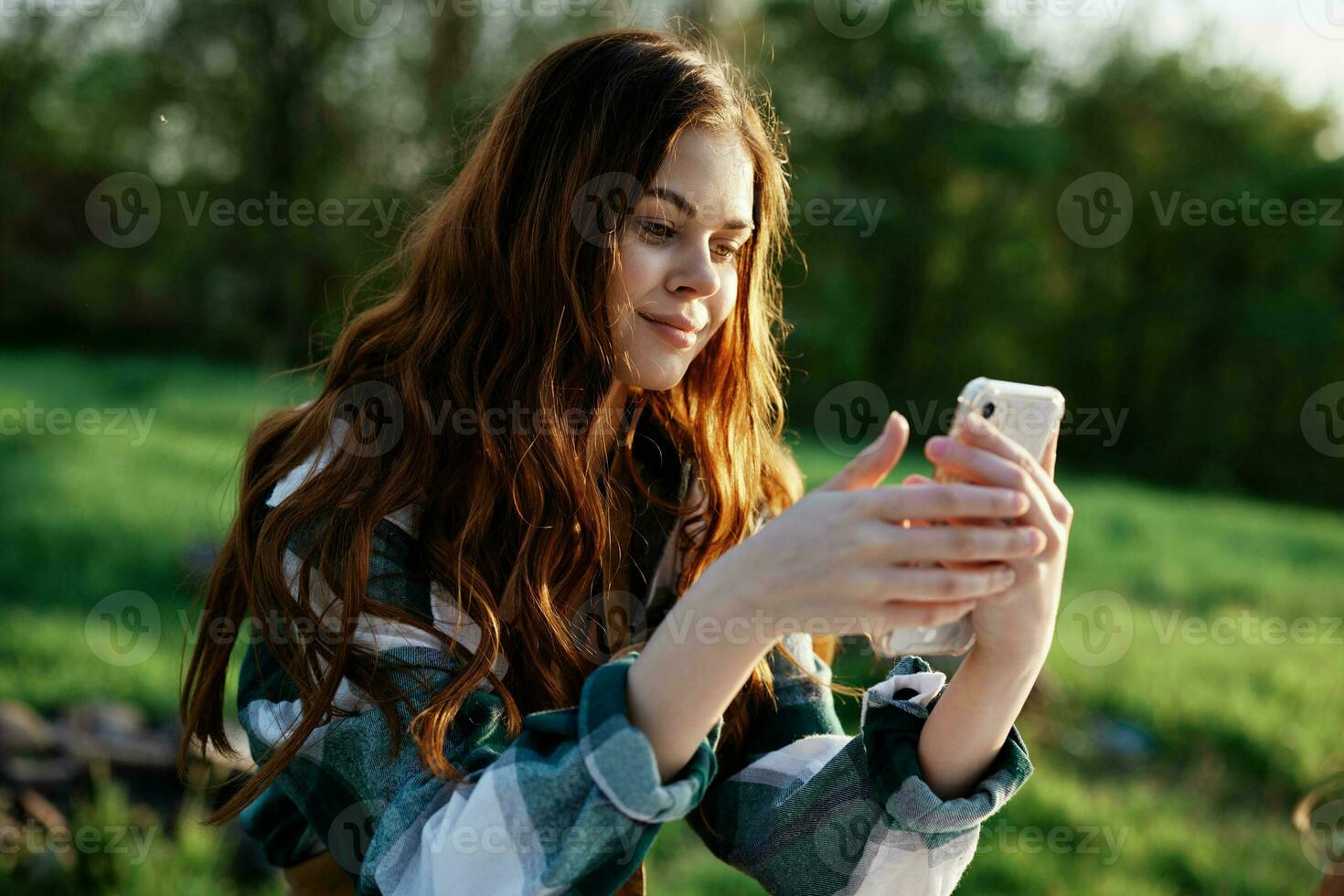 en skön kvinna avkopplande och arbetssätt på henne telefon Sammanträde i natur i de parkera bland de träd leende och innehav henne smartphone i henne hand belyst förbi de ljus solnedgång ljus foto