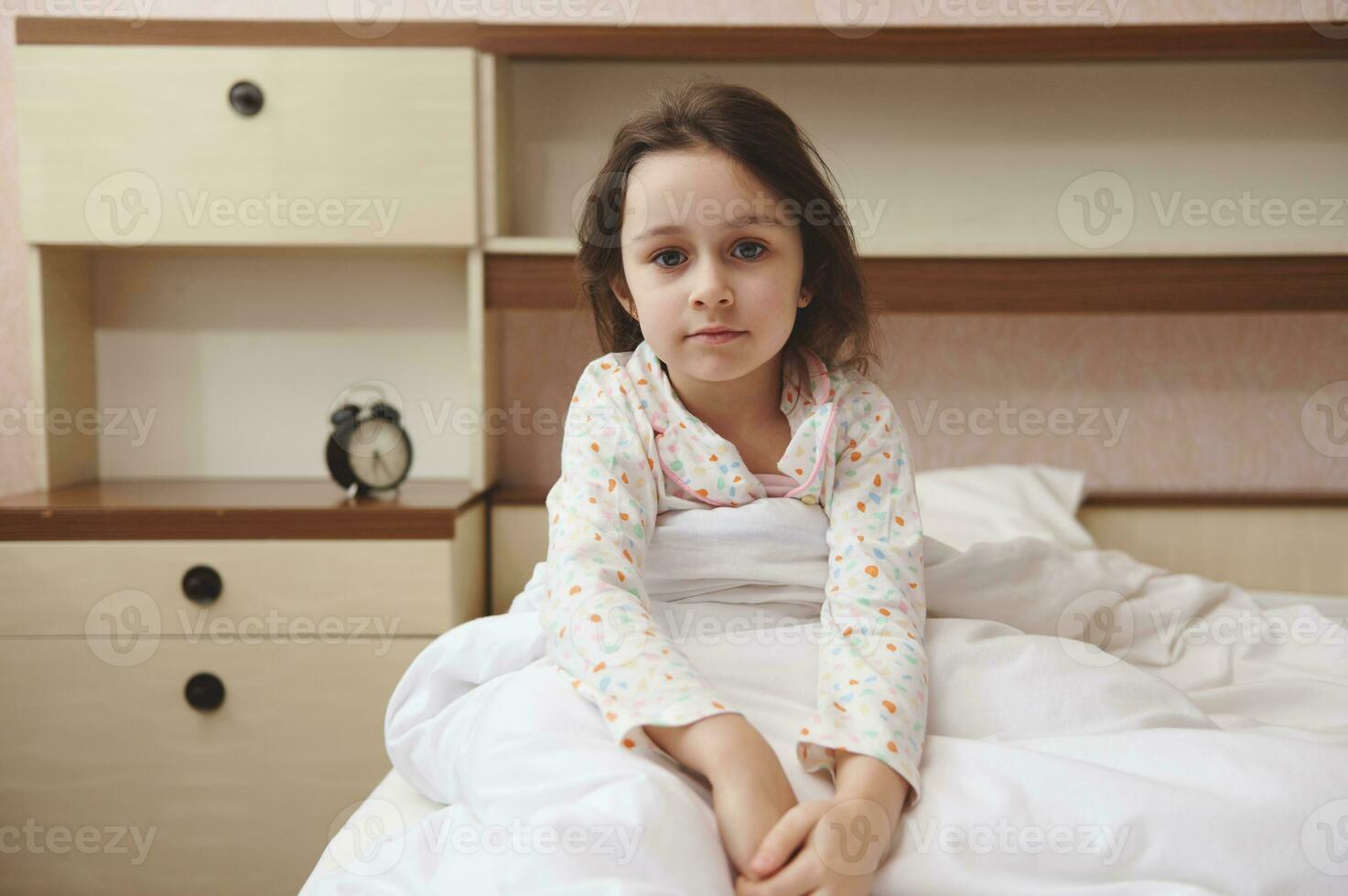 porträtt av en caucasian skön barn flicka i pyjamas, Sammanträde i de säng på henne mysigt sovrum foto