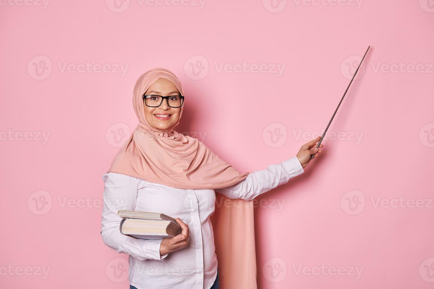 gravid muslim kvinna lärare i rosa hijab och glasögon, leende på kamera, pekande på kopia Plats, förklara lektion foto