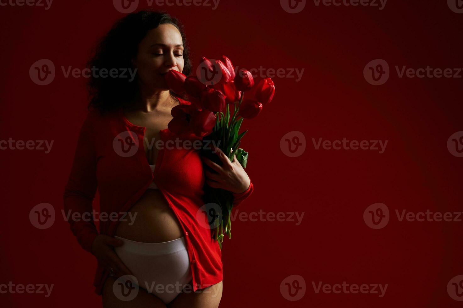 underbar latin amerikan vuxen gravid kvinna smeksam henne mage, sniffa en söt bukett av röd tulpaner för mors dag foto