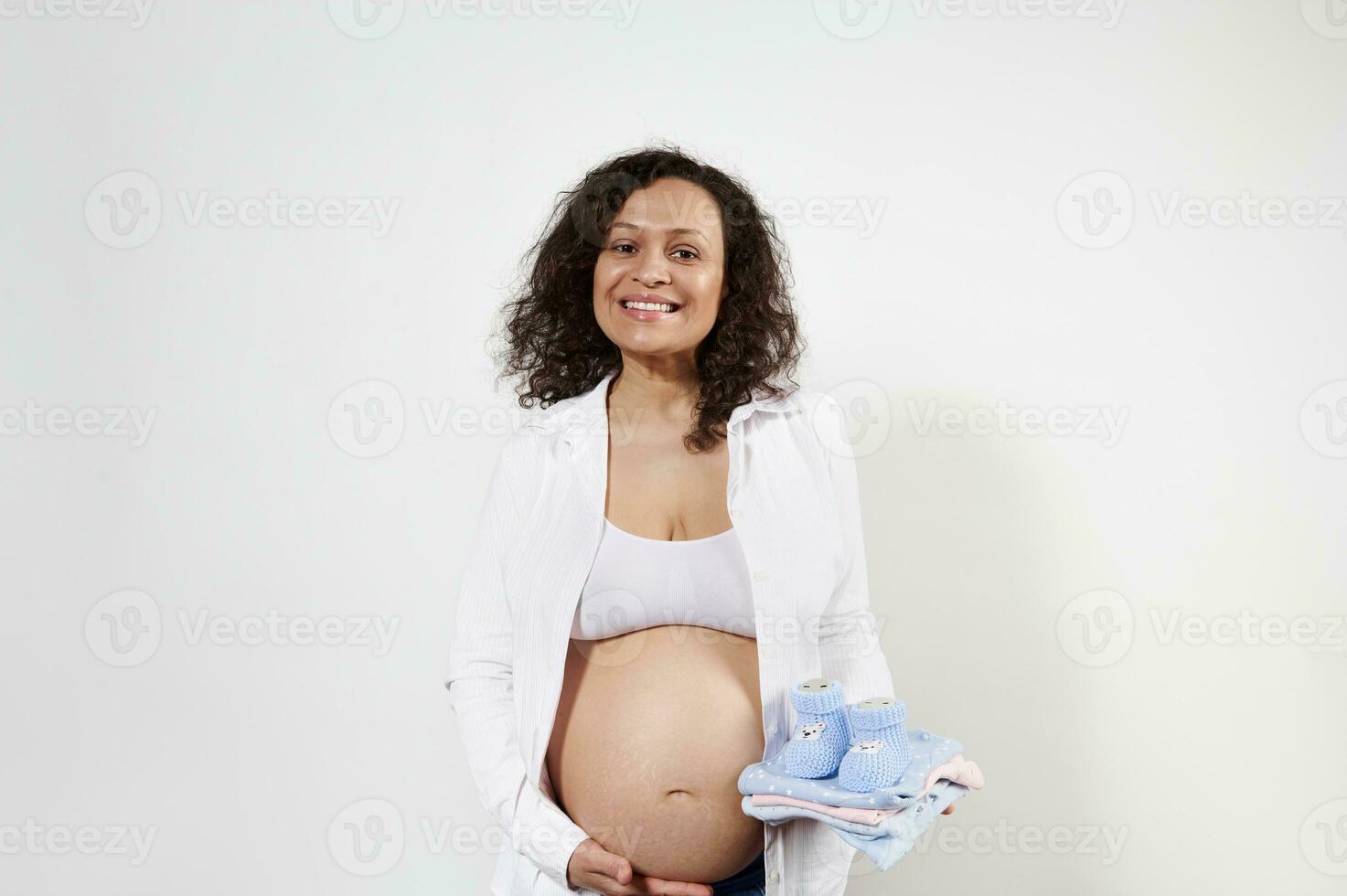 gravid kvinna försiktigt rörande henne mage, leende på kamera, Framställ med blå stickat bebis tossor på vit bakgrund. foto