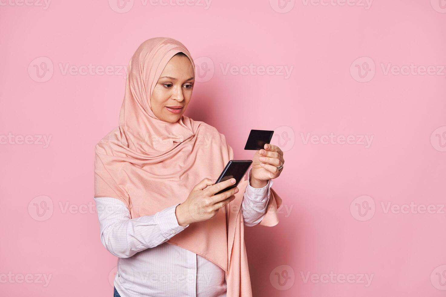 Lycklig gravid muslim kvinna i hijab, innehav kreditera kort och smartphone. mobil appar. internet bankverksamhet. uppkopplad handla foto