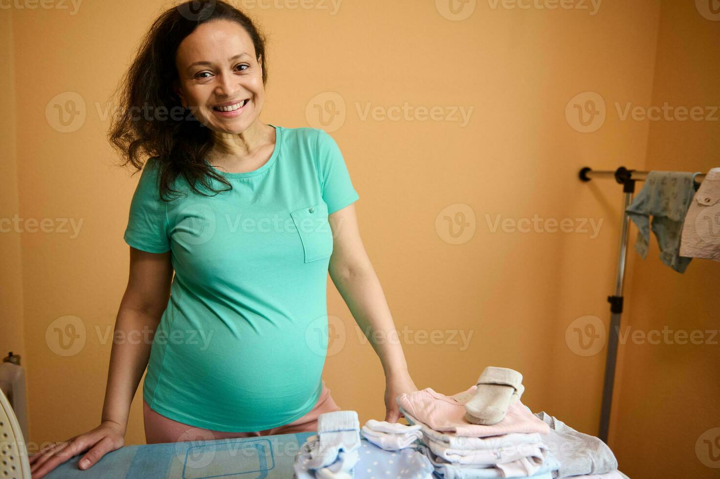 gravid kvinna med positiv känsla och skön leende på henne ansikte, stående förbi strykning styrelse med strykas bebis kläder foto