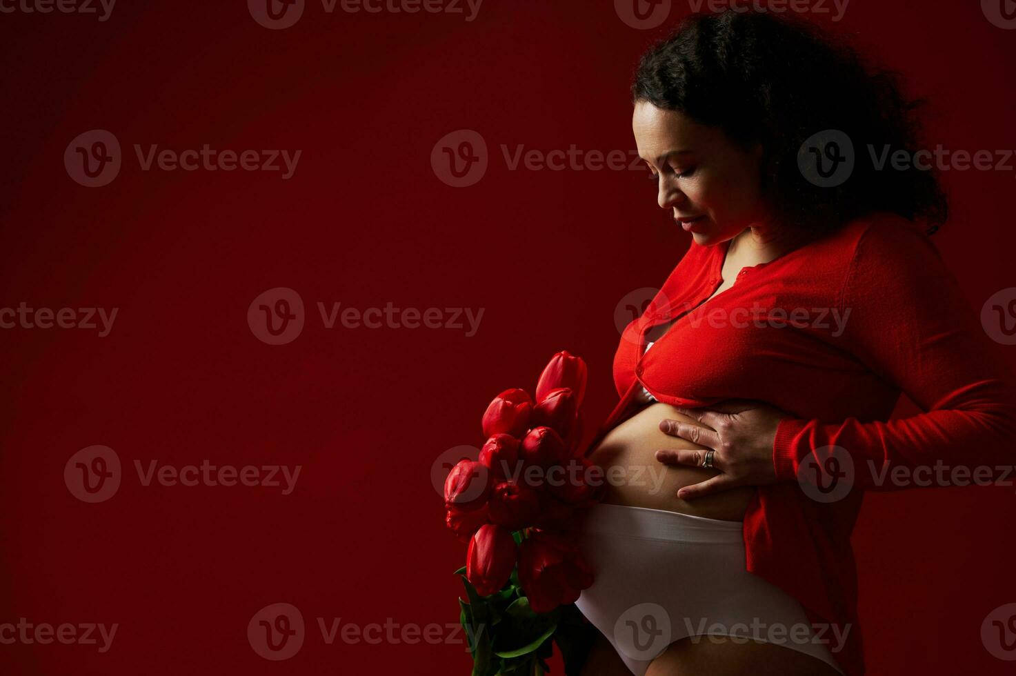 äkta porträtt av skön gravid kvinna innehav en bukett av röd tulpaner och smeksam henne mage på röd bakgrund foto