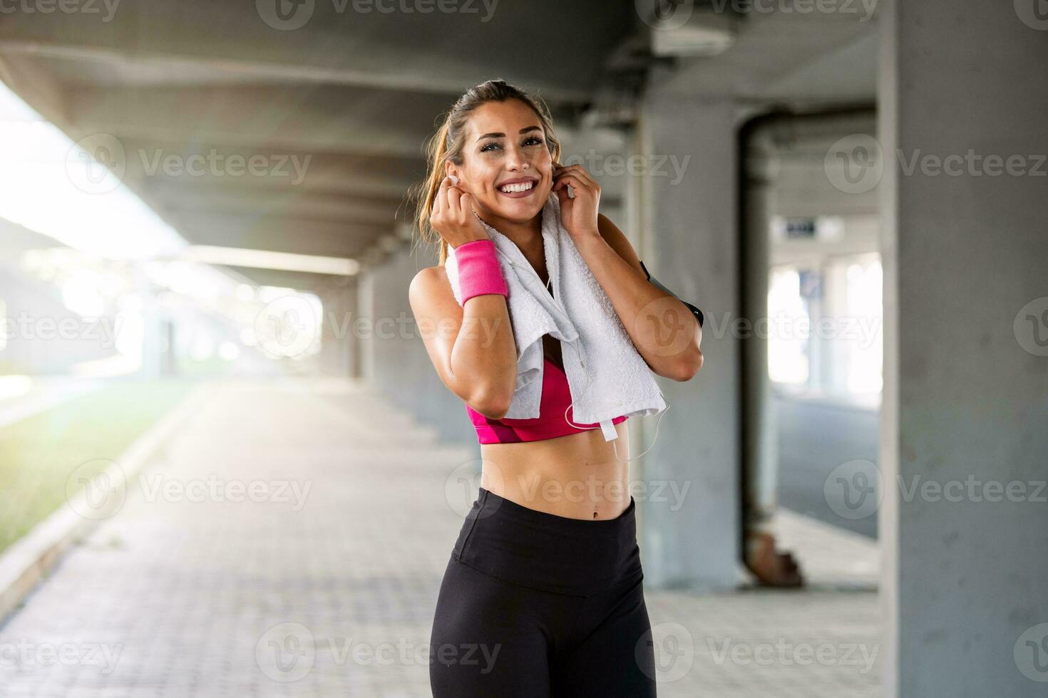 porträtt av förtjusande charmig leende ung form kondition flicka med hörlurar innehav en handduk och Framställ. kondition kvinna tar en ha sönder efter löpning träna. foto