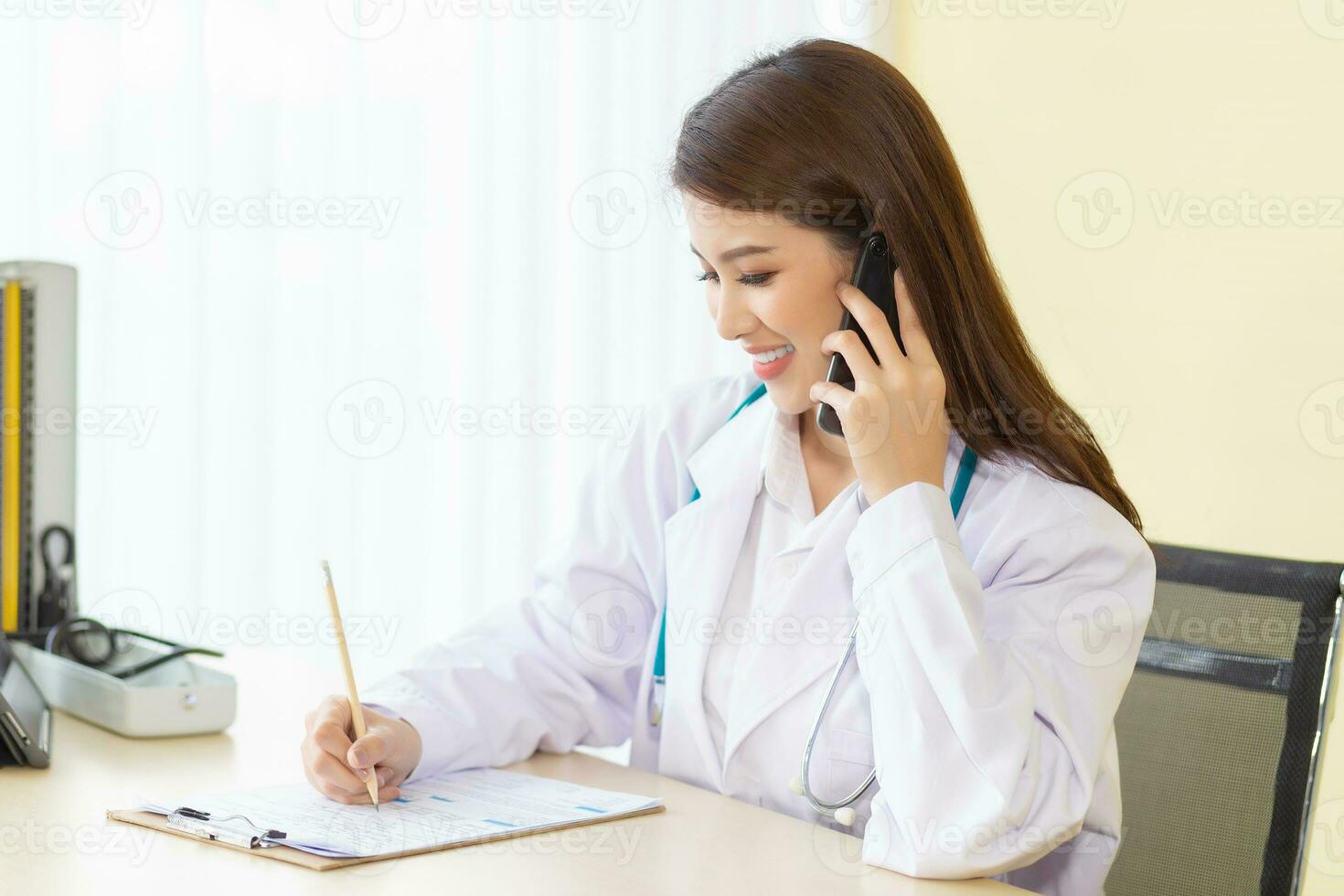 professionell ung asiatisk kvinna läkare bär en vit morgonrock och stetoskop Sammanträde med använda sig av smartphones till prata fråga Följ upp på patienter symptom i de undersökning rum på sjukhus. foto