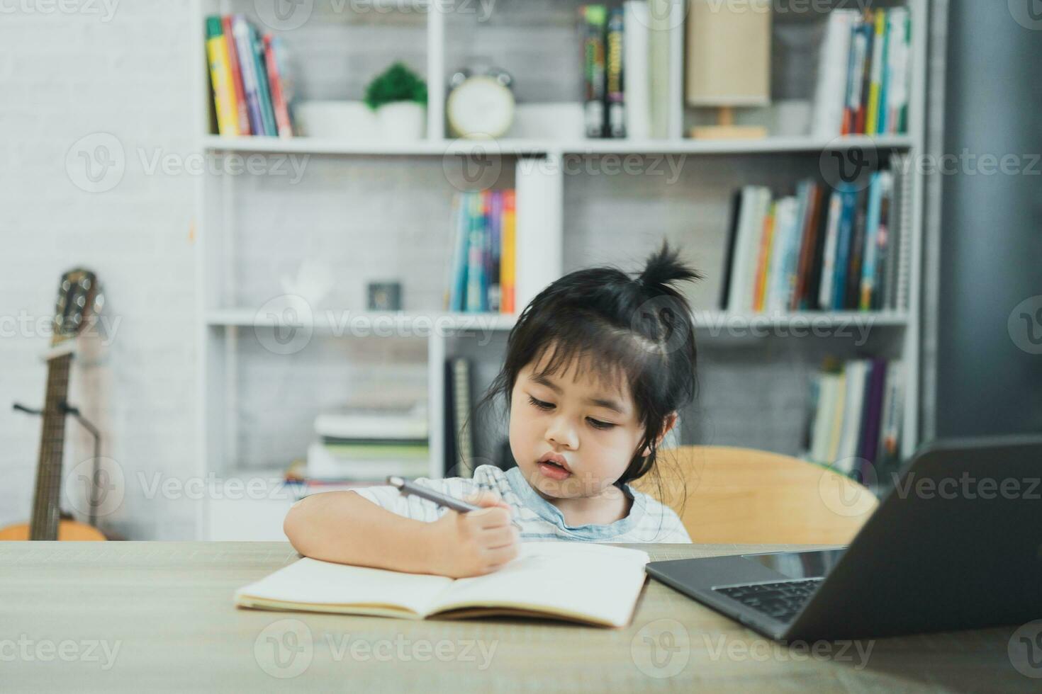 asiatisk bebis flicka bär en blå randig skjorta använda sig av bärbar dator och skriva anteckningar i anteckningsbok till studie uppkopplad på trä tabell skrivbord i levande rum på Hem. utbildning inlärning uppkopplad från Hem begrepp. foto