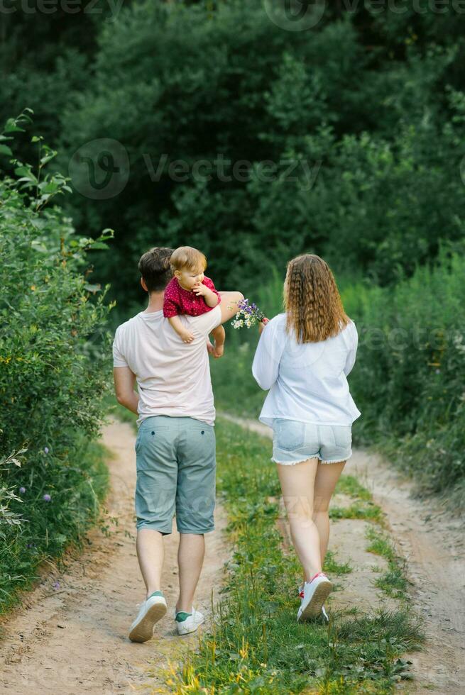 Lycklig ung familj med deras son i deras vapen är gående längs en skog väg och njuter de sommar väder foto