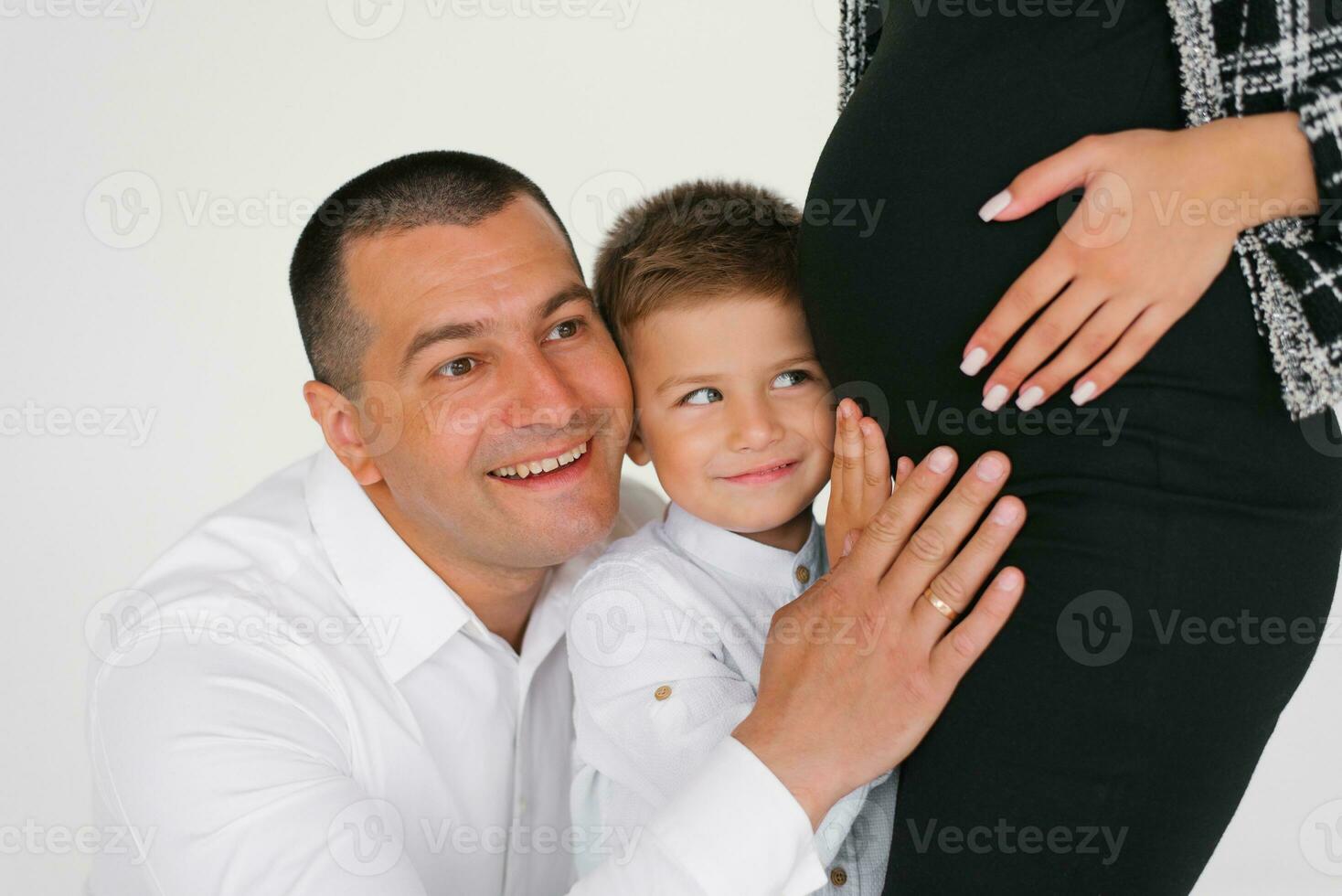 Lycklig bebis pojke, son och pappa lyssnar och innehar hans händer på hans gravid mors mage medan väntar för en bror eller syster foto