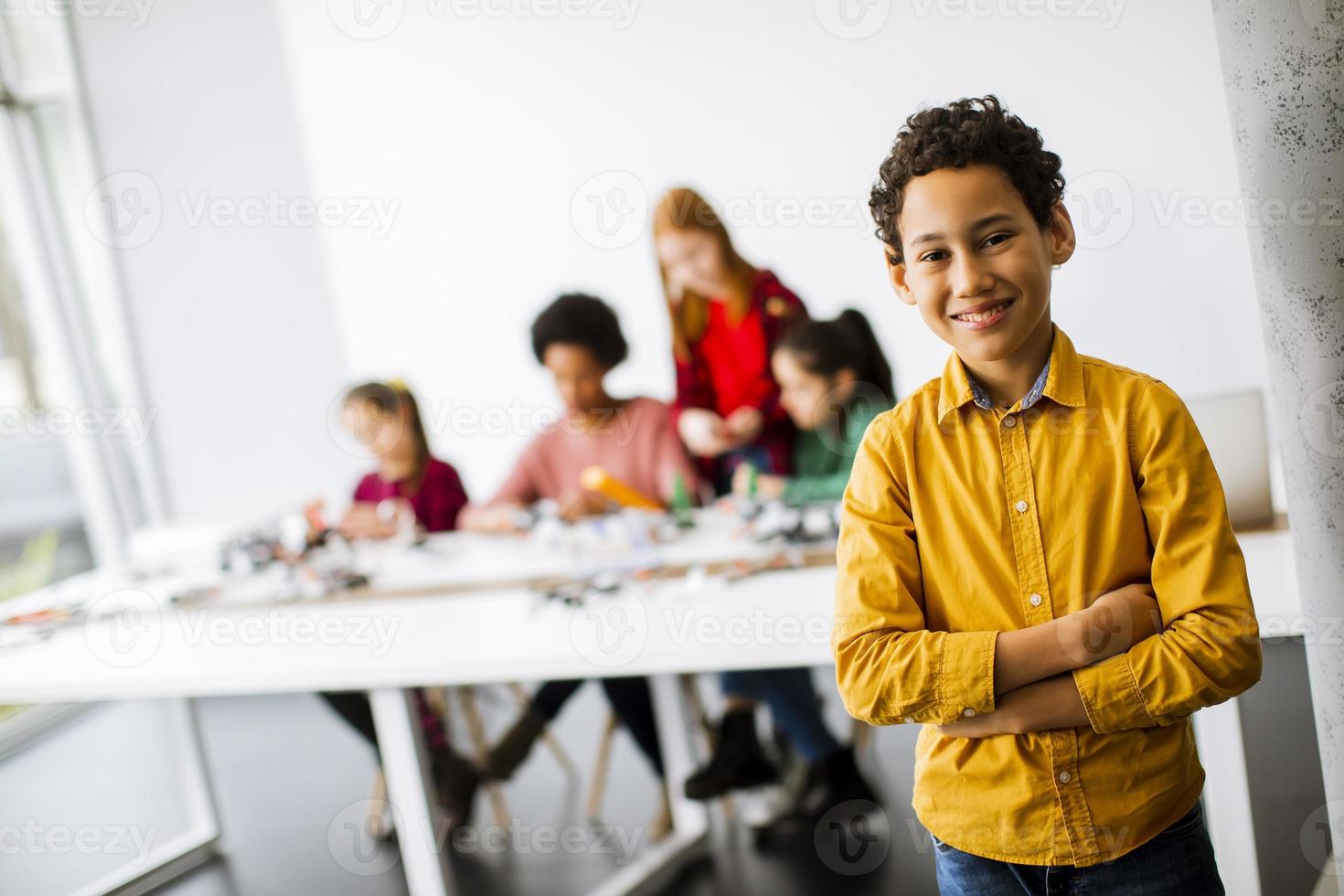 söt liten pojke som står framför barn som programmerar elektriska leksaker och robotar i klassrummet för robotik foto