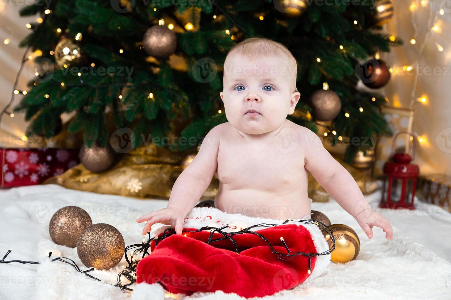 bedårande bebis utan kläder på jultomtenhatt på en bakgrund av julbollar foto