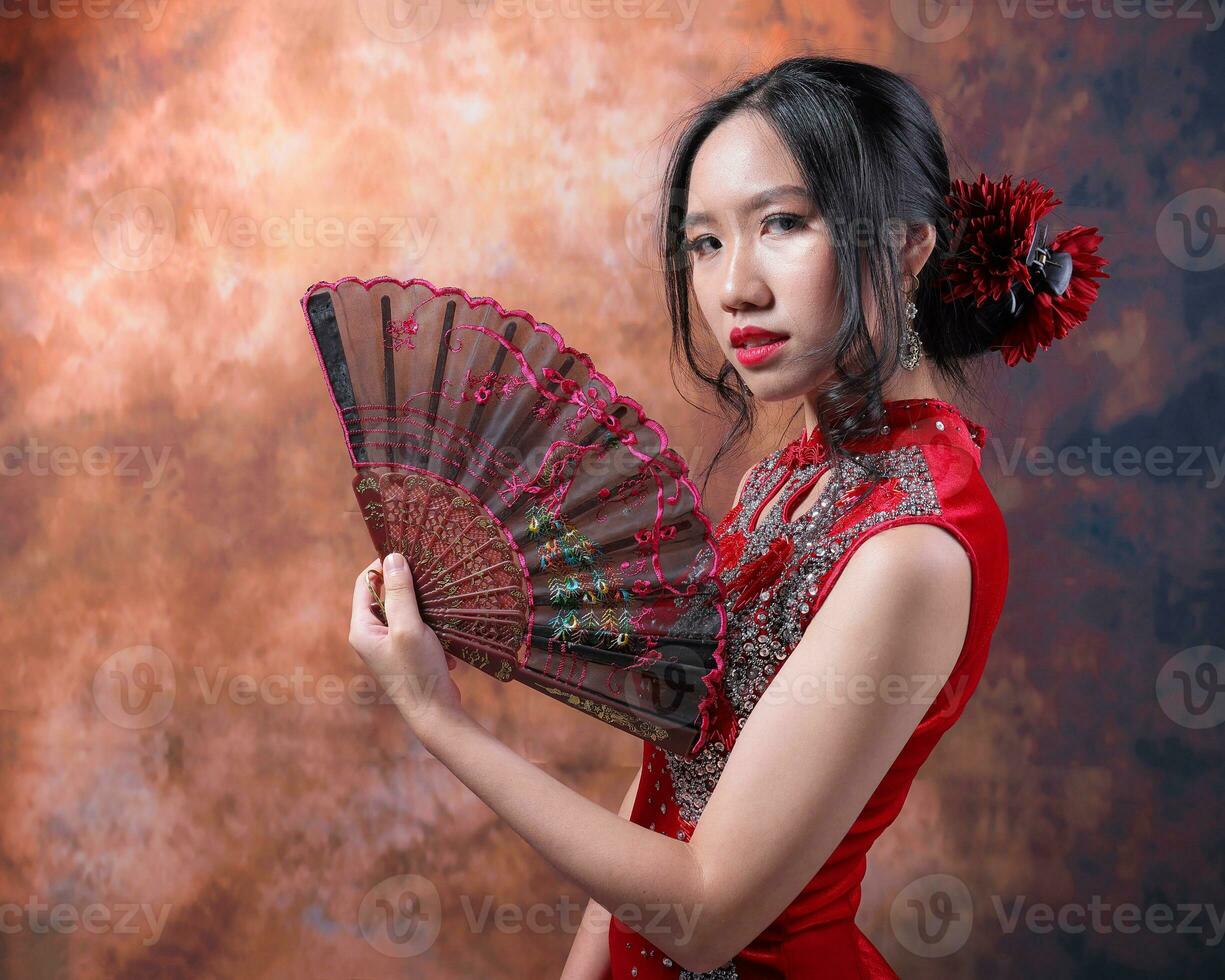 söder öst asiatisk kinesisk lopp etnisk ursprung kvinna bär röd sammet Cheongsam med hand sys sekvens arbete klänning kostym hand fläkt på retro årgång bakgrund foto