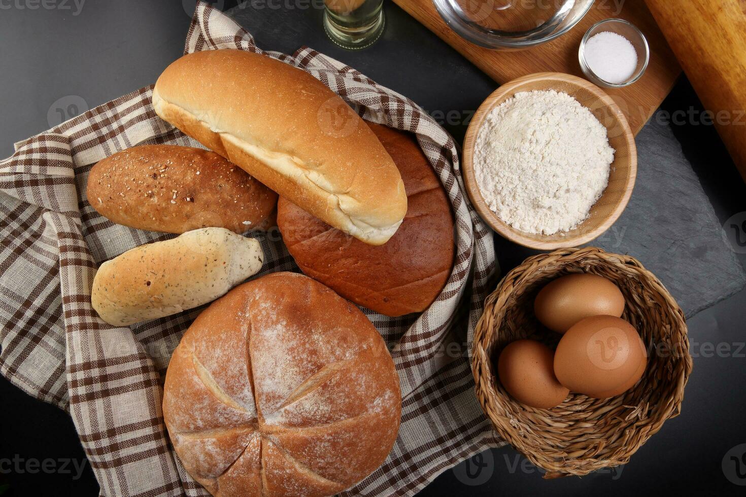 nyligen bakad bröd limpa bulle rulla runda lång blanda sanning insvept i rutig kök tyg servett handduk vete blomma olja vatten salt ägg rullande stift skiffer sten över svart bakgrund foto