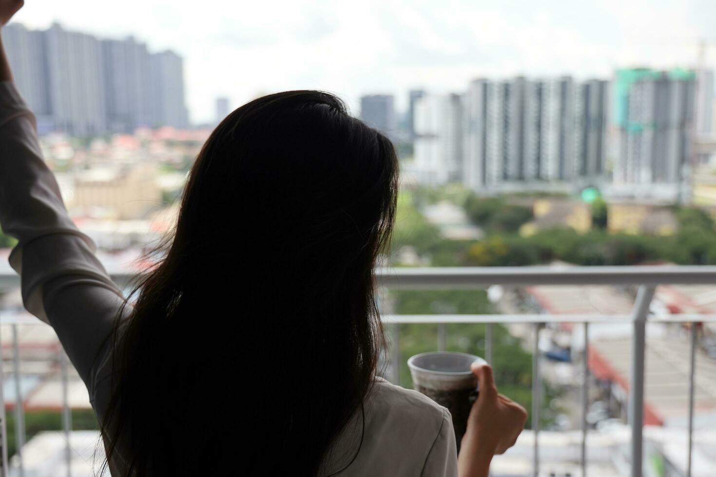 skön ung sydöst asiatisk kvinna innehav te kaffe kopp nära balkong fönster stad scape bakgrund, ser avkopplande tänkande foto