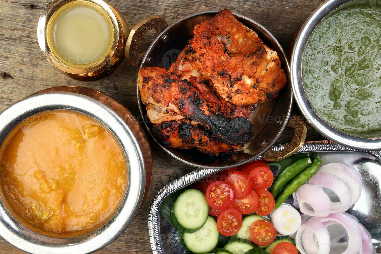 kryddad röd kyckling grillad tikka tandoori nan bröd grön spenat curry yoghurt sås dal tomat gurka lök sallad uppsättning i metall rostfri stål mässing koppar pott på rustik årgång trä bakgrund foto