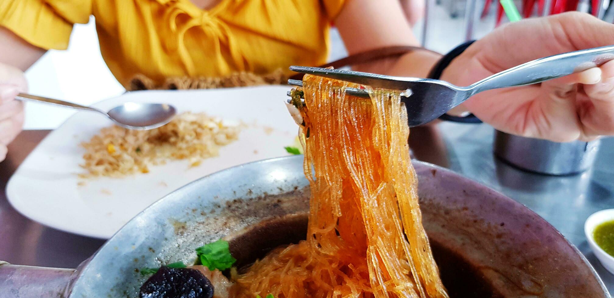 närbild äter glas spaghetti med gaffel och kvinna i gul skjorta eller klänning suddig bakgrund på lokal- mat restaurang. äter thai eller Asien mat på lunch eller frukost tid. foto
