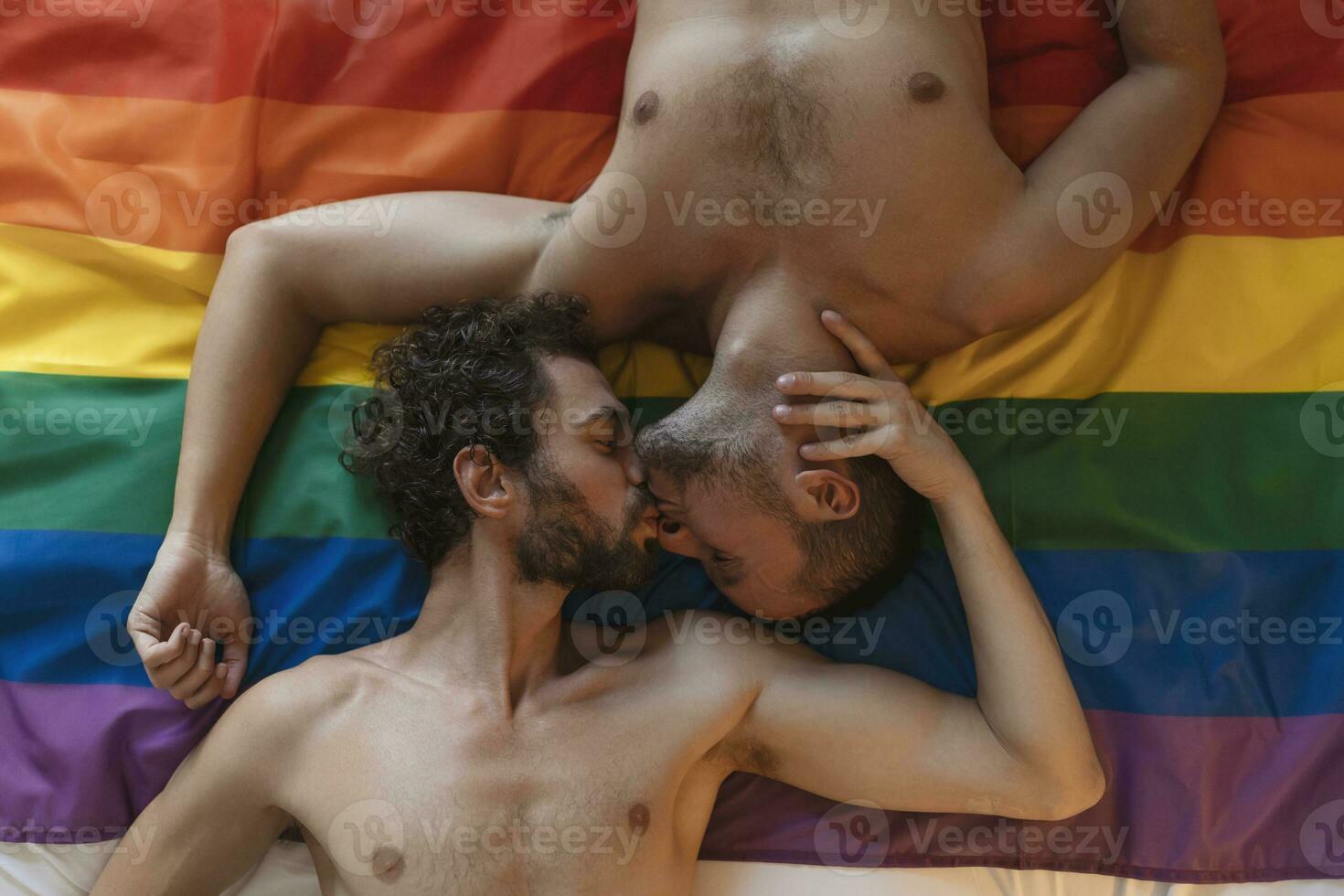 öm ung Gay man kissing hans älskare på de säng. två ung manlig älskande om tillsammans på stolthet flagga. romantisk ung Gay par bindning förtjust inomhus. foto