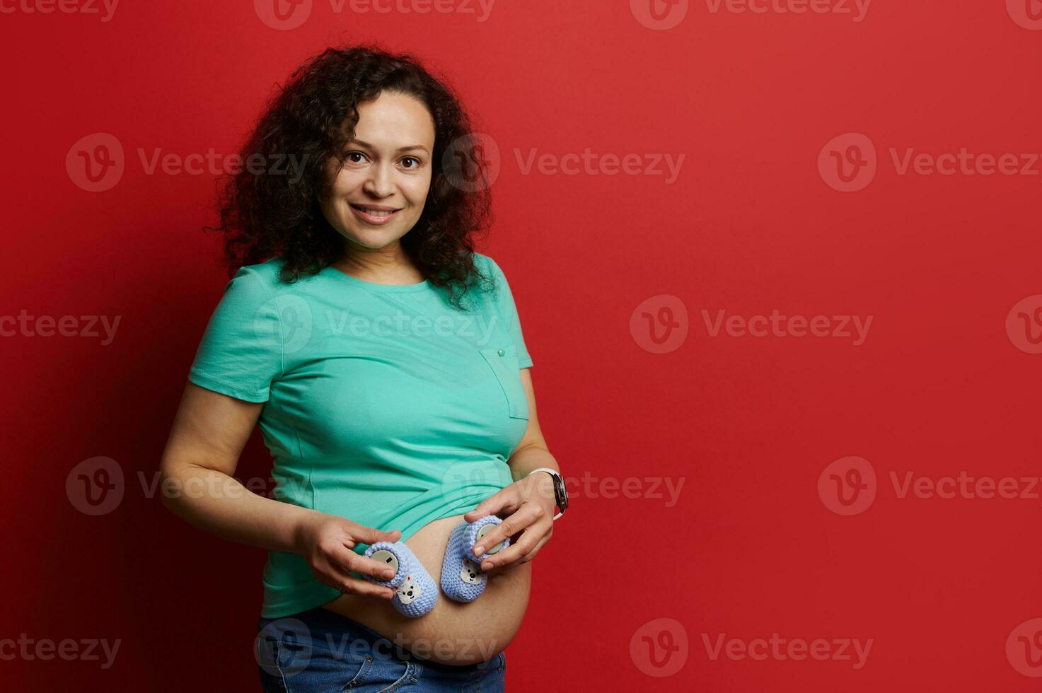 Lycklig gravid kvinna försiktigt rörande mage, leende på kamera, Framställ med blå stickat bebis tossor över röd bakgrund foto