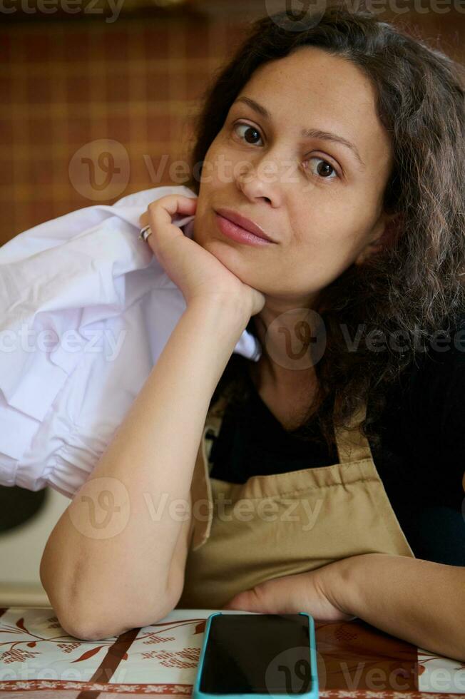 närbild porträtt av en självsäker skön kvinna, kock konditor i beige förkläde, innehav vit keps, ser på kamera foto