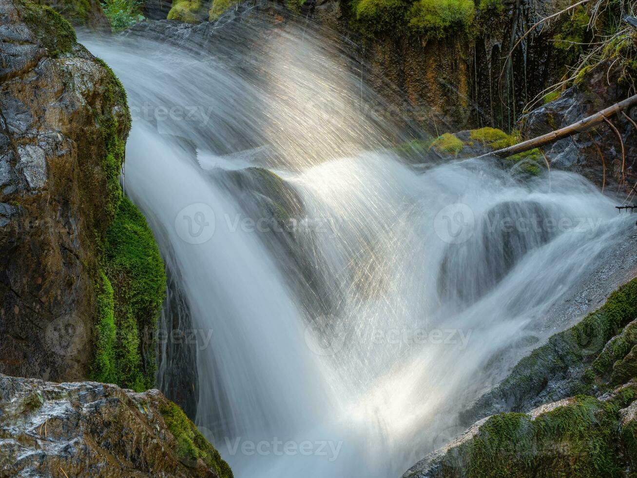 kaskader av en vattenfall i en grön berg skog stänga upp. natur bakgrund av turbulent faller vatten ström på våt rocks. vatten jets är upplyst förbi de Sol. foto