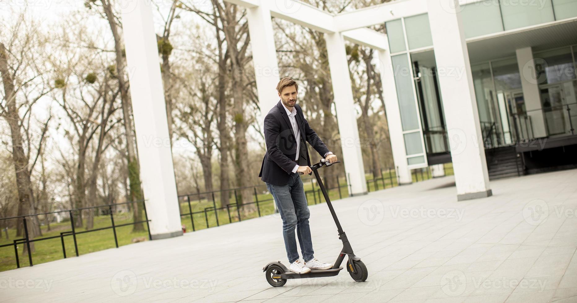 ung affärsman i avslappnade kläder som rider på en elektrisk skoter vid en kontorsbyggnad på ett affärsmöte foto