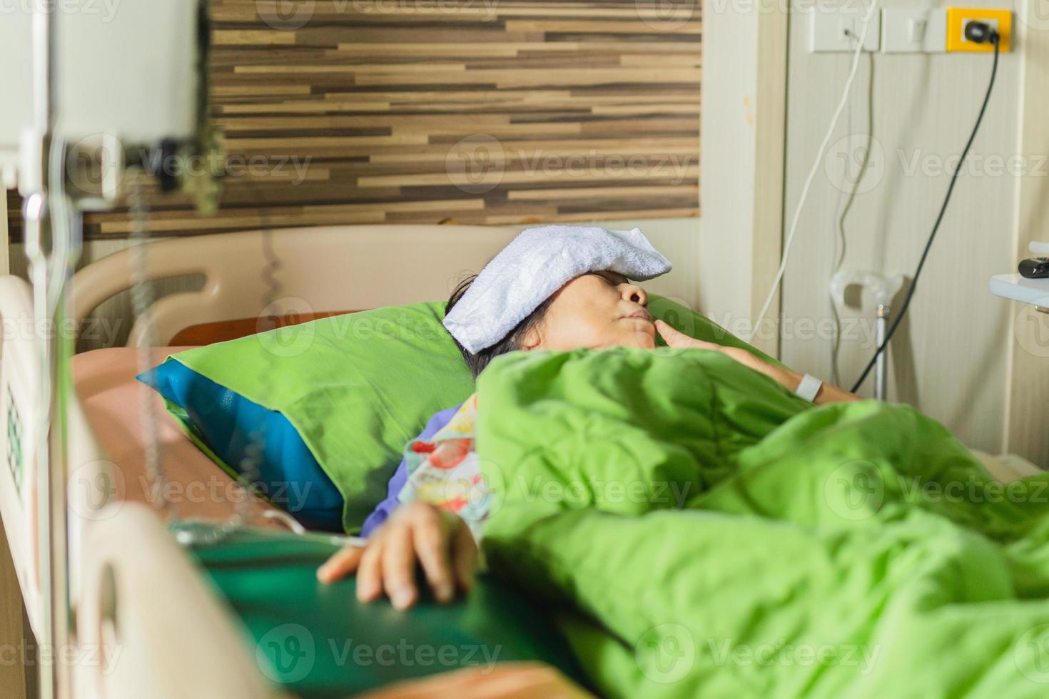 äldre kvinnapatient som ligger i sängen på sjukhus med kompress på pannan foto