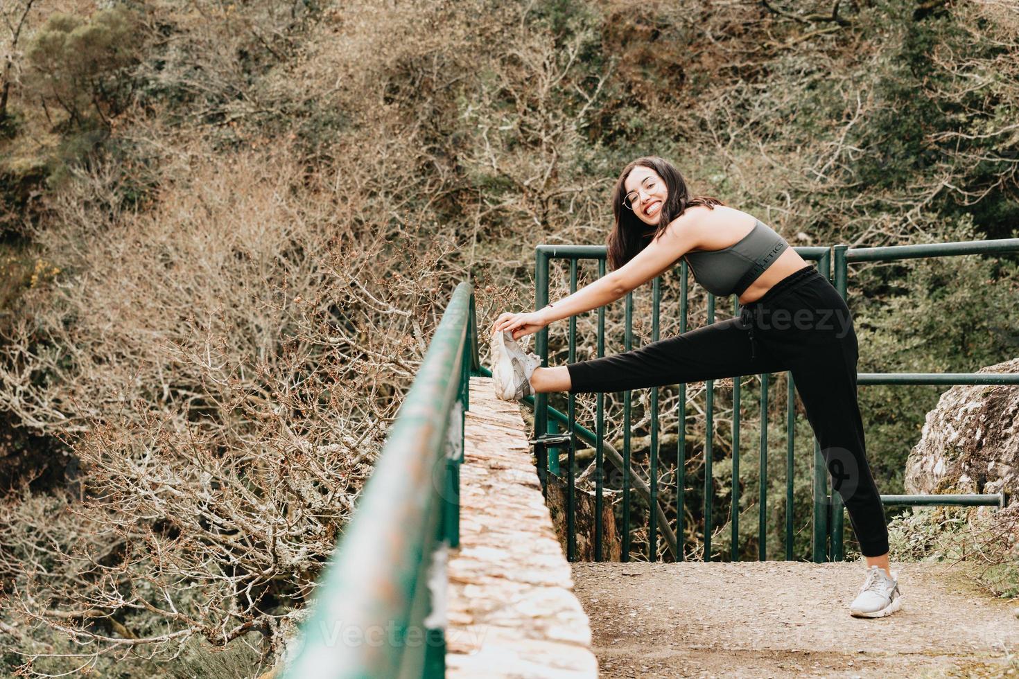 ung kvinna på sportkläder som sträcker benet över en trottoar mitt i en höstlig skog medan du ler mot kameran kopiera utrymme fitness koncept foto