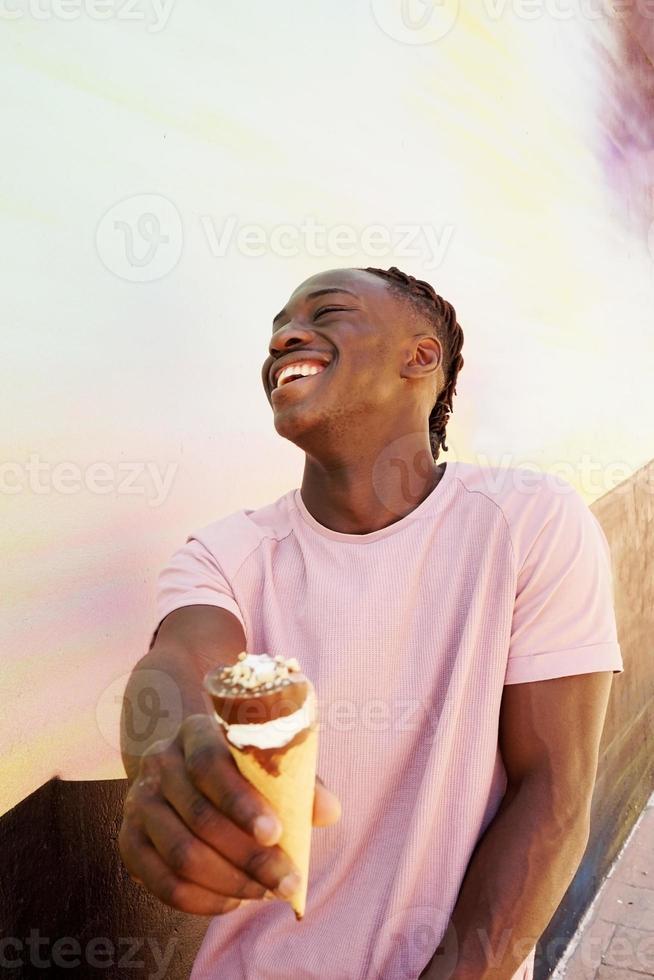 ung stilig svart man bär en rosa skjorta håller och äter en glasskotte på sommaren på en målad vägg som soluppgång eller solig dag foto