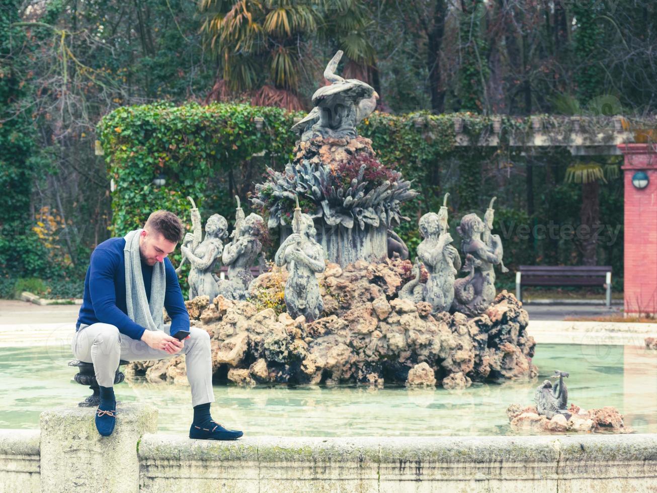 ung man sitter i fontänen i en park och tittar på sin mobiltelefon foto