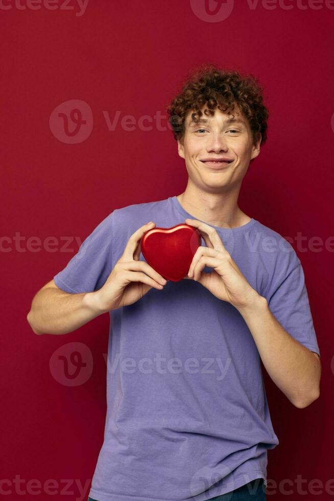 rödhårig lockigt kille en låda i de form av en hjärta i hans händer röd bakgrund foto