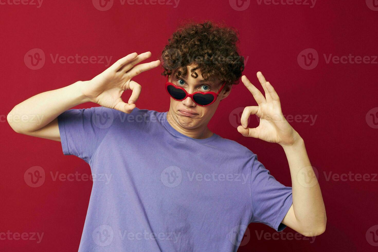 kille med röd lockigt hår kille i en lila t-shirt med solglasögon Framställ röd bakgrund foto