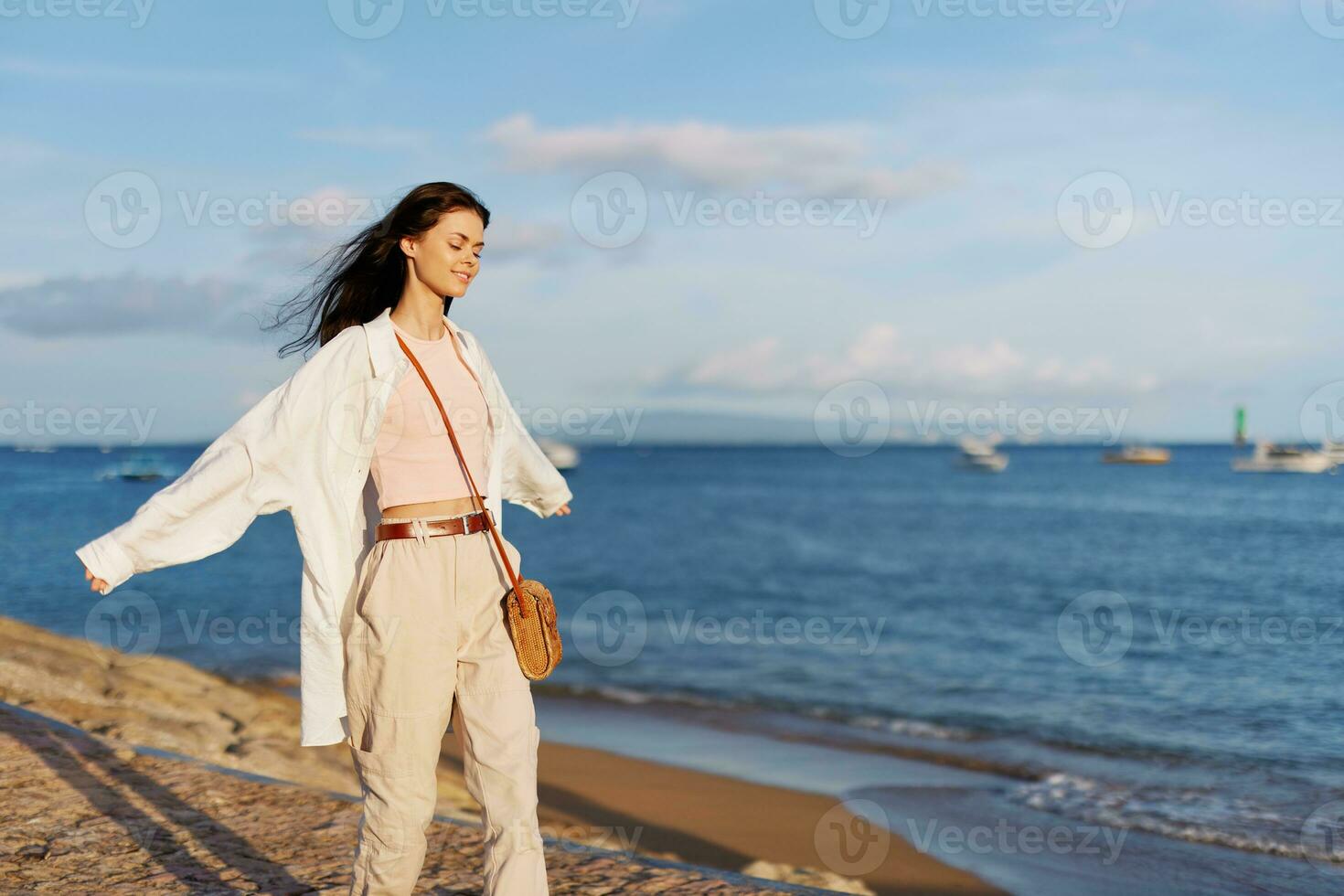 en kvinna Lycklig med henne ögon stängd i de Sol gående på de strand mot de bakgrund av de hav leende med tänder, flygande hår, solbränd hud, avslappning, en resa till de hav och frihet. foto